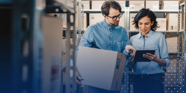 two coworkers standing ina. warehouse reading items off of a tablet while one of them holds a large box