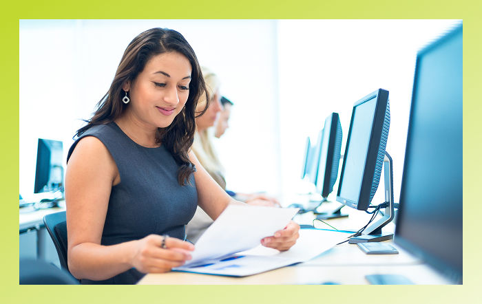 Business person smiling and reviewing papers at a desk, in front of a computer